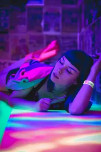 A woman with dark bangs lies on a colorful, glowing surface in a room bathed in purple and pink light, representing a virtual performance.
